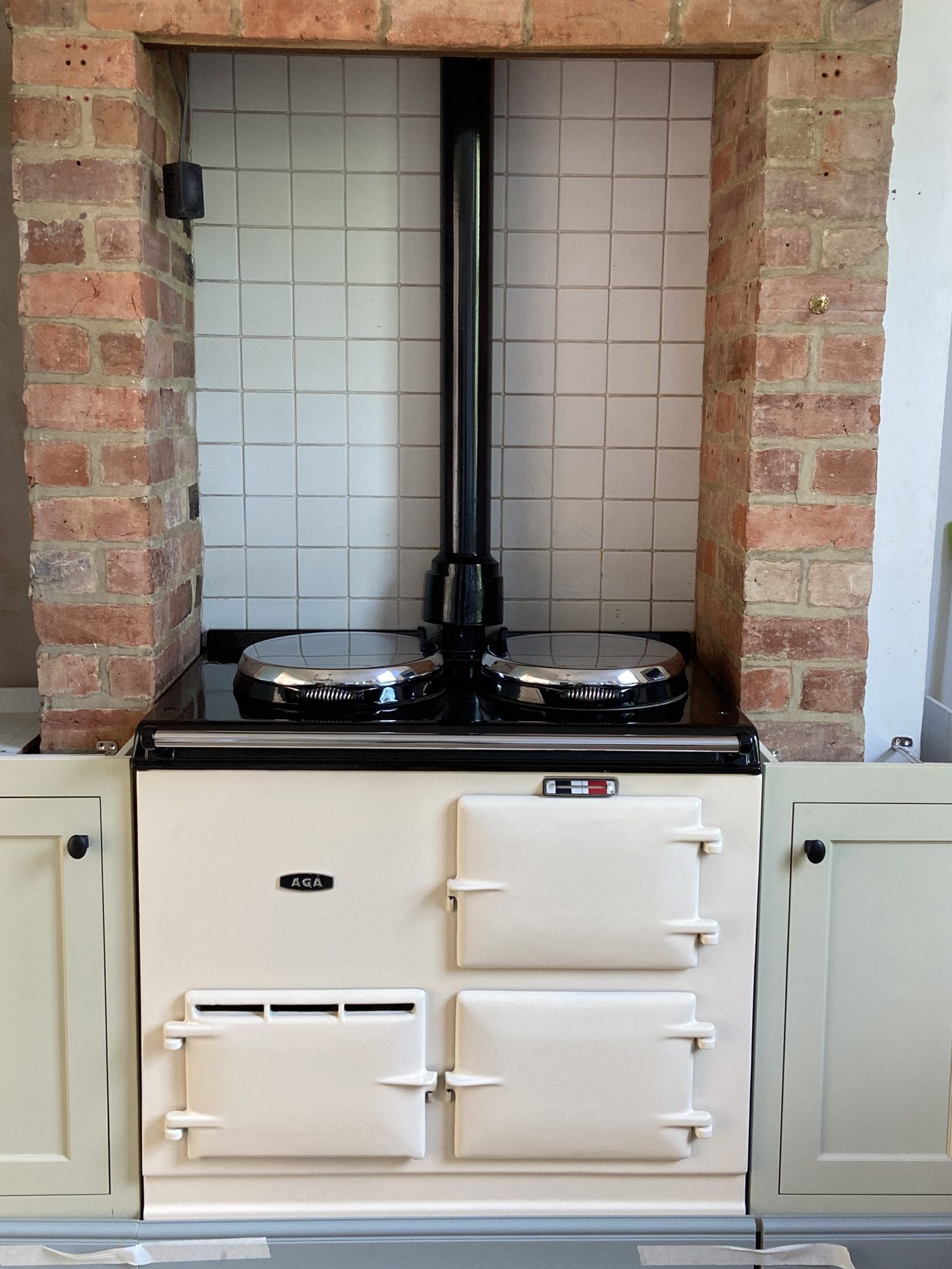 A freshly re-enamelled cream two-oven AGA in a brick fireplace surround, between sage green kitchen units
