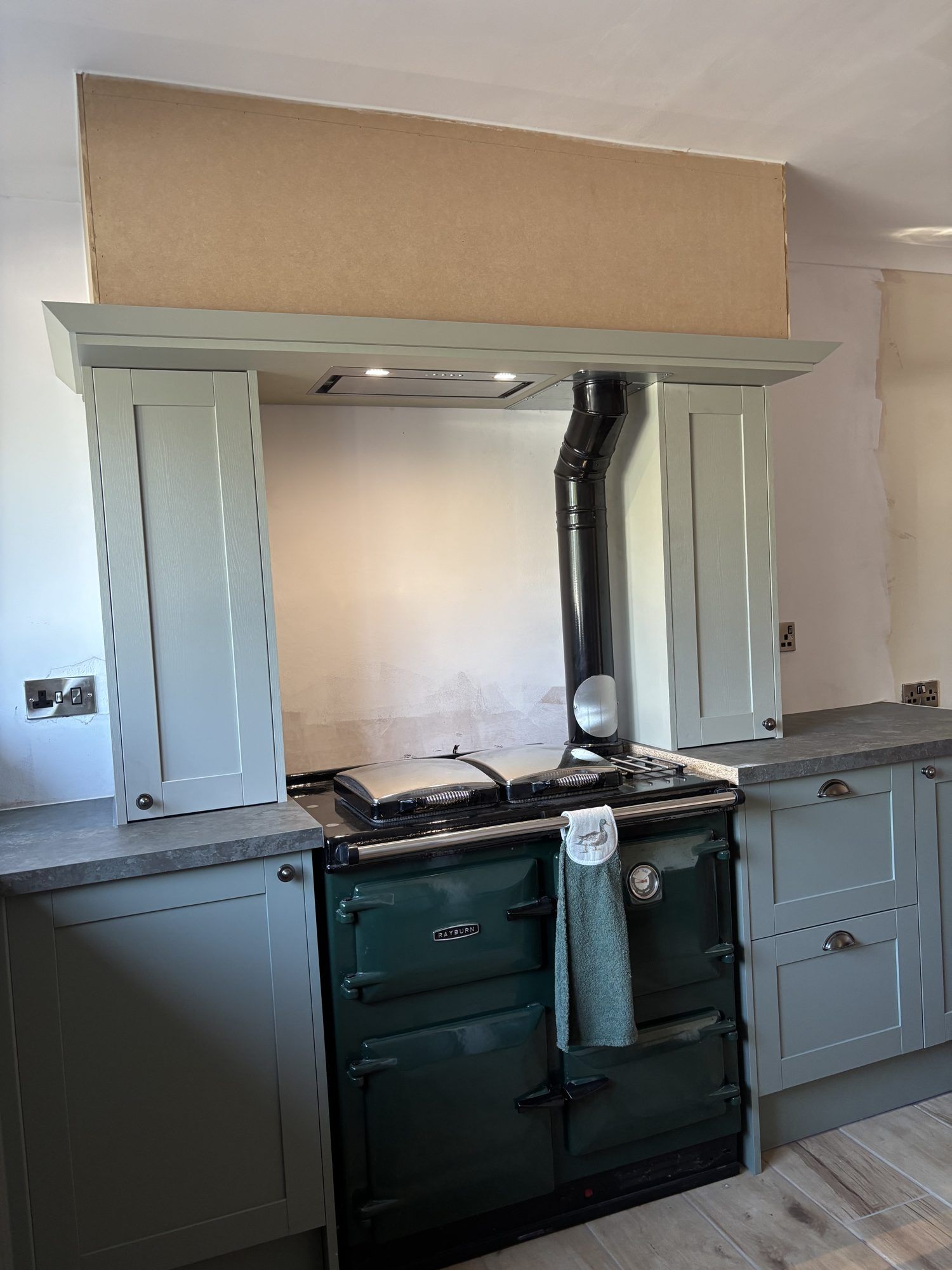 A green gas Rayburn fitted between sage green kitchen cabinets, with its black flue pipe rising behind the hotplate lids