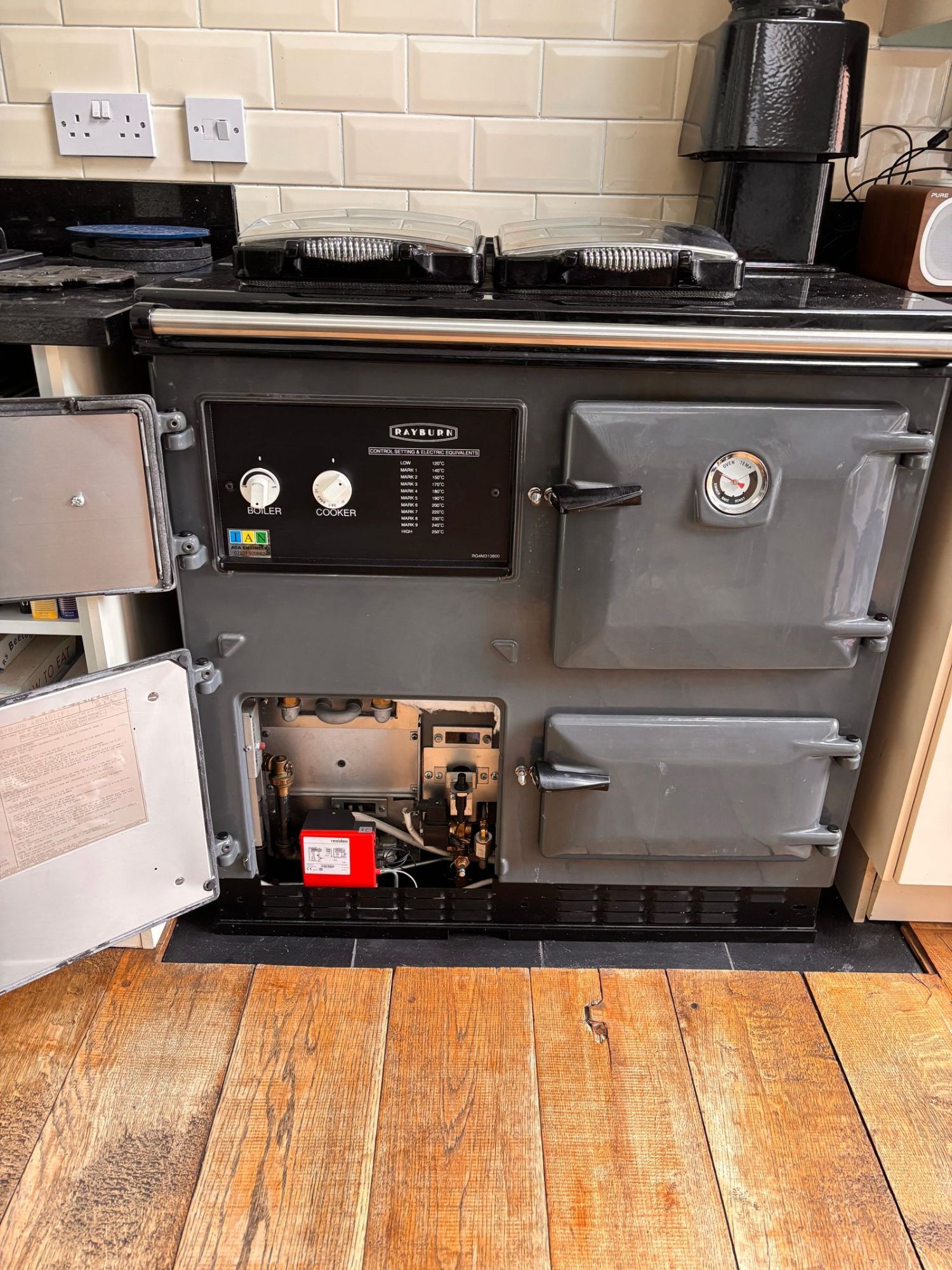 A grey gas Rayburn with its burner compartment doors open, showing the gas valve, pipework and the control panel with the boiler and cooker dials