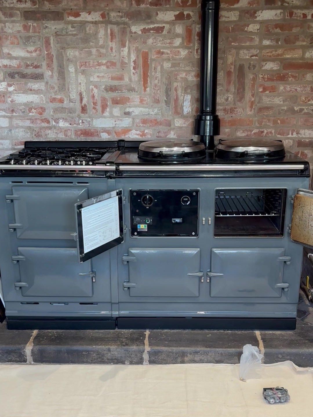 A grey AGA with an integrated module and gas hob against a brick chimney breast, its control panel exposed, oven doors open and a gas valve on the dust sheet in front