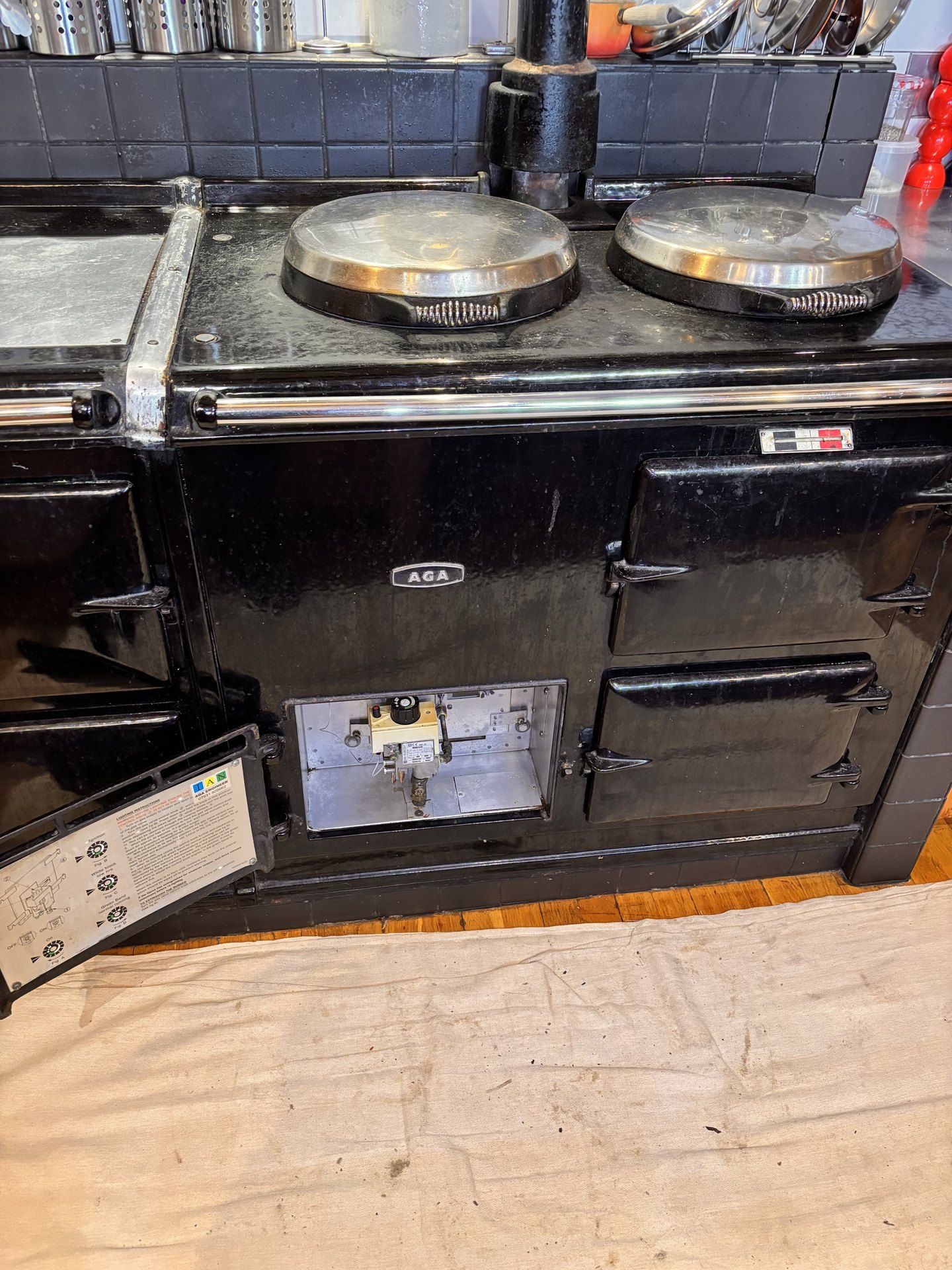 A black AGA with its burner door open, showing the gas valve and controls inside, above a dust sheet on a wooden floor