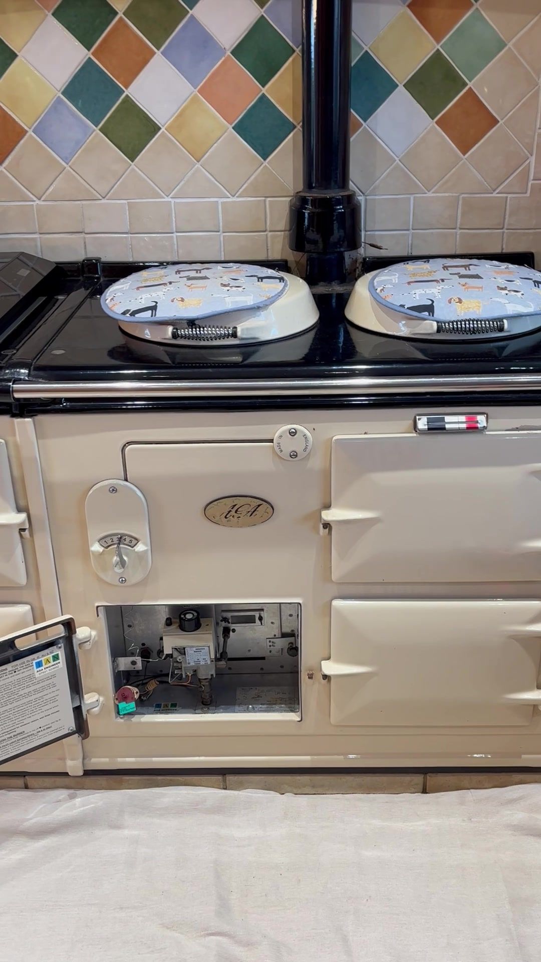 A cream gas AGA with its burner door open, showing the gas valve and controls, under a black flue pipe and a wall of coloured diamond tiles