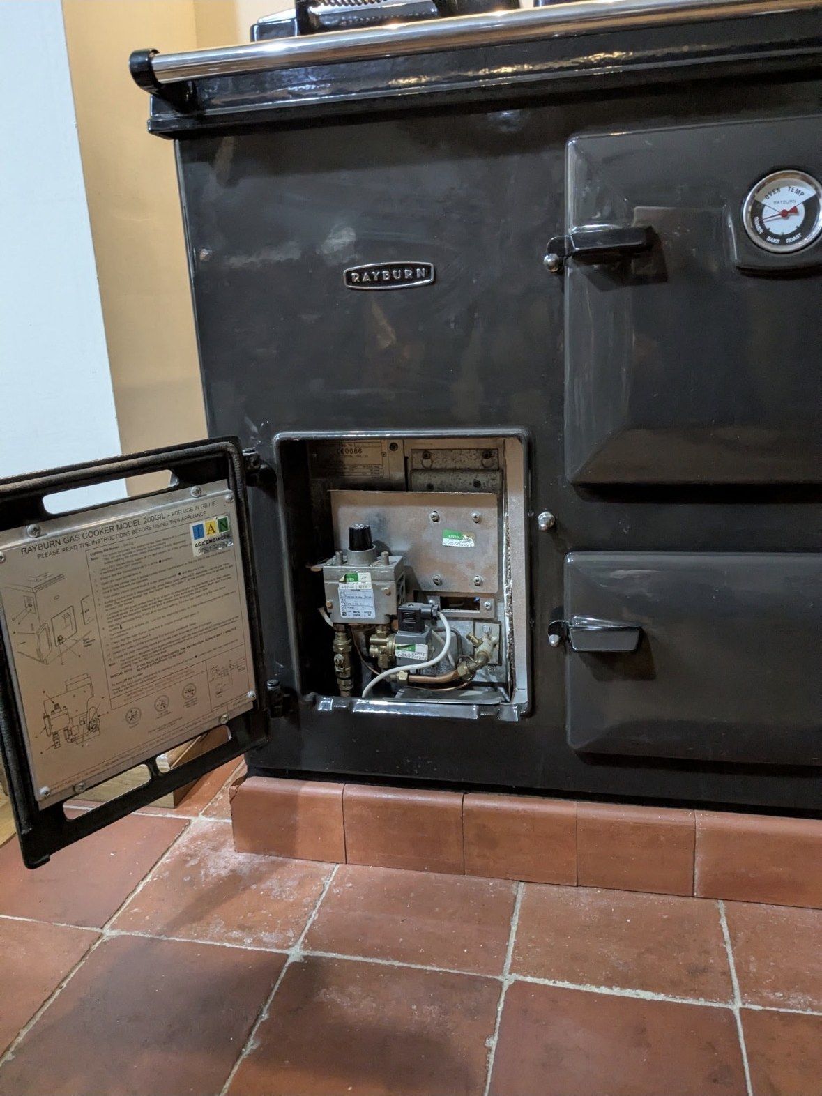 A black gas Rayburn 200G with its burner door open, showing the gas valve and pipework, on a quarry-tiled floor