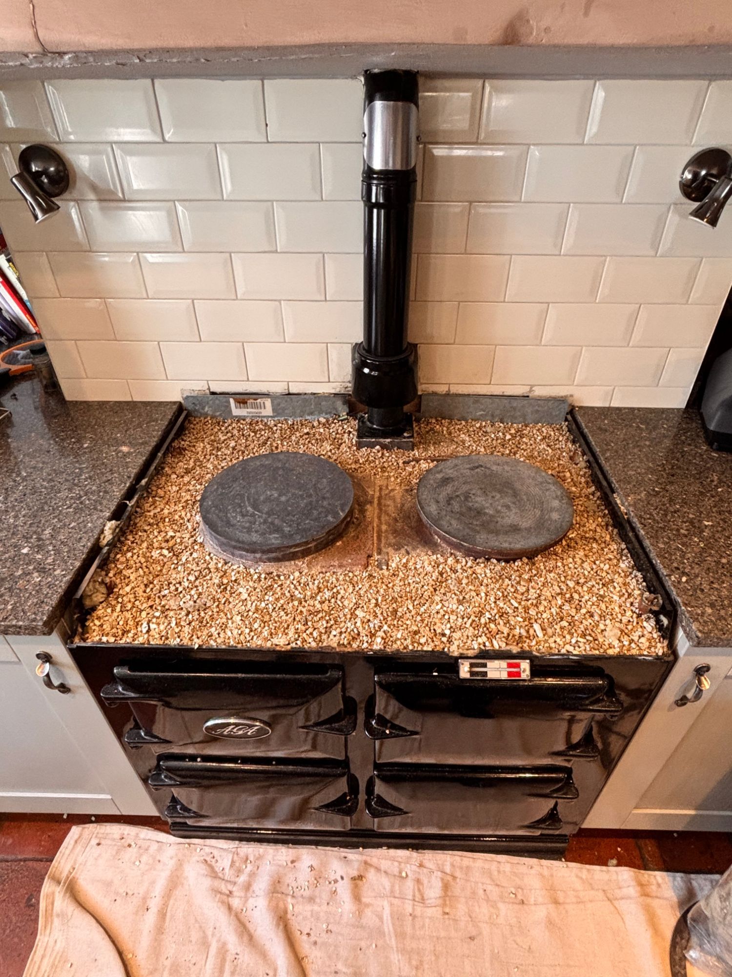 A black two-oven AGA with its top plate off, showing the two hotplates sitting in loose insulation granules, under a black flue pipe and white brick tiles