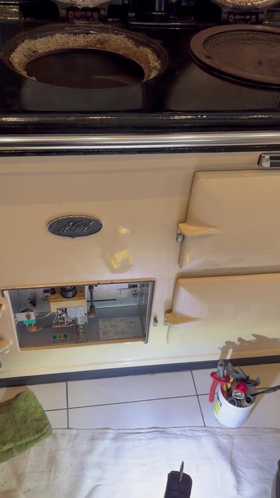 A cream gas AGA with its burner door open to show the gas valve and controls, and the left hotplate lifted out to show the insulation around it
