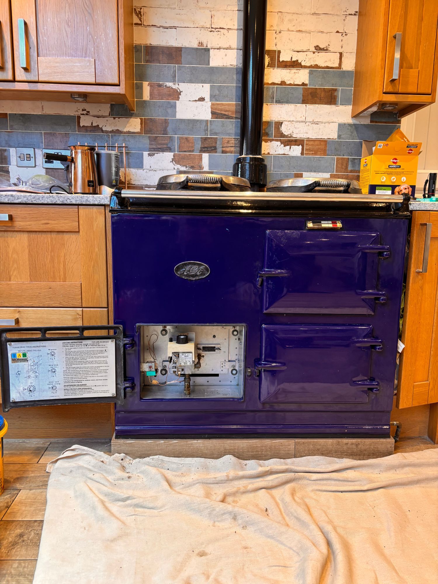 A dark blue two-oven gas AGA in an oak kitchen with its burner door open to show the gas valve and controls, and a dust sheet on the floor in front