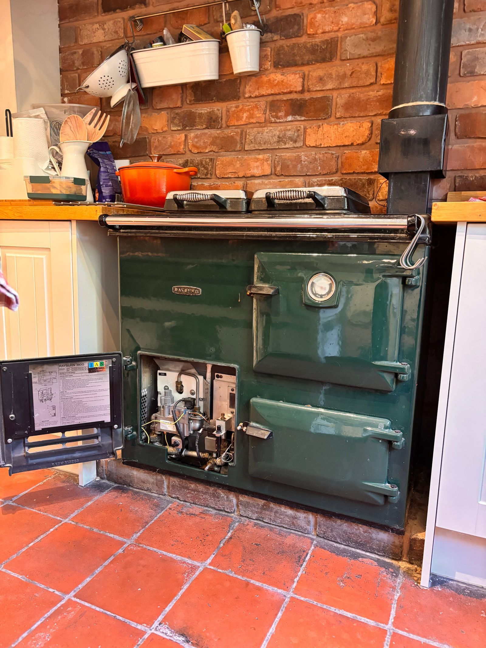 A dark green gas Rayburn 380G with its burner door open to show the gas valve and controls, against a brick wall on a quarry-tiled floor