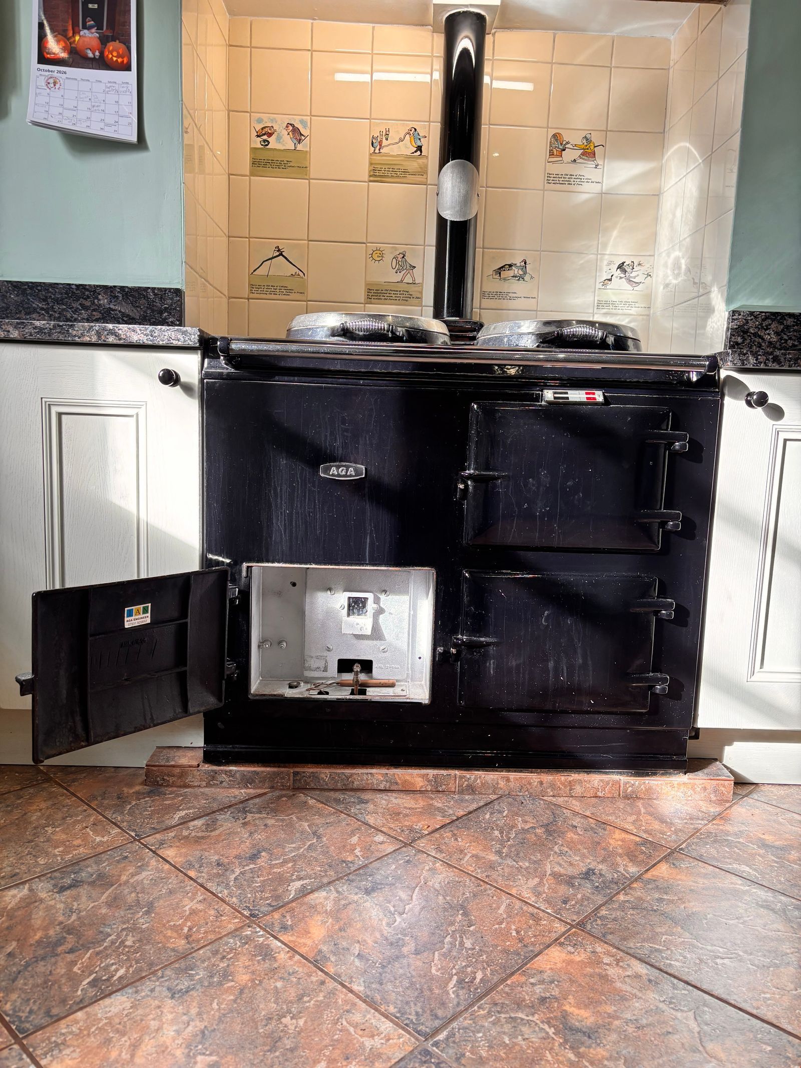 A black two-oven oil AGA with its burner door open, under a tiled alcove with a black flue pipe, on a tiled kitchen floor