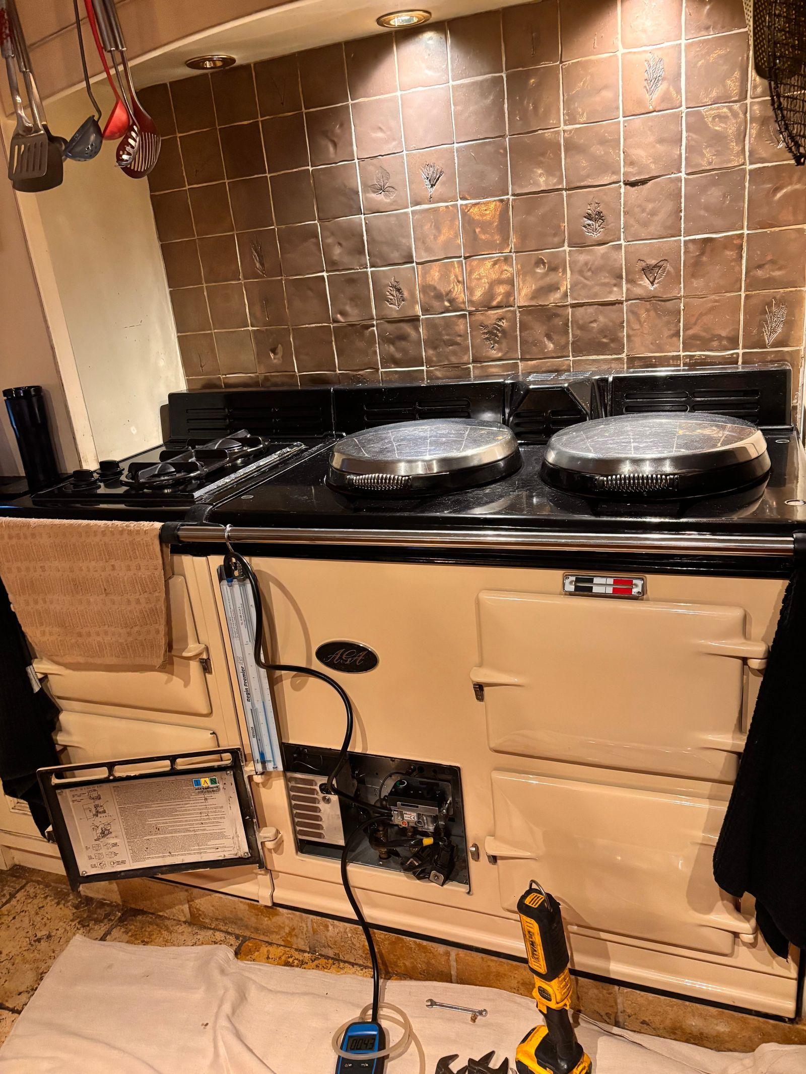 A cream gas AGA with its burner door open and pressure gauges connected, in front of bronze tiles, with a dust sheet on the floor