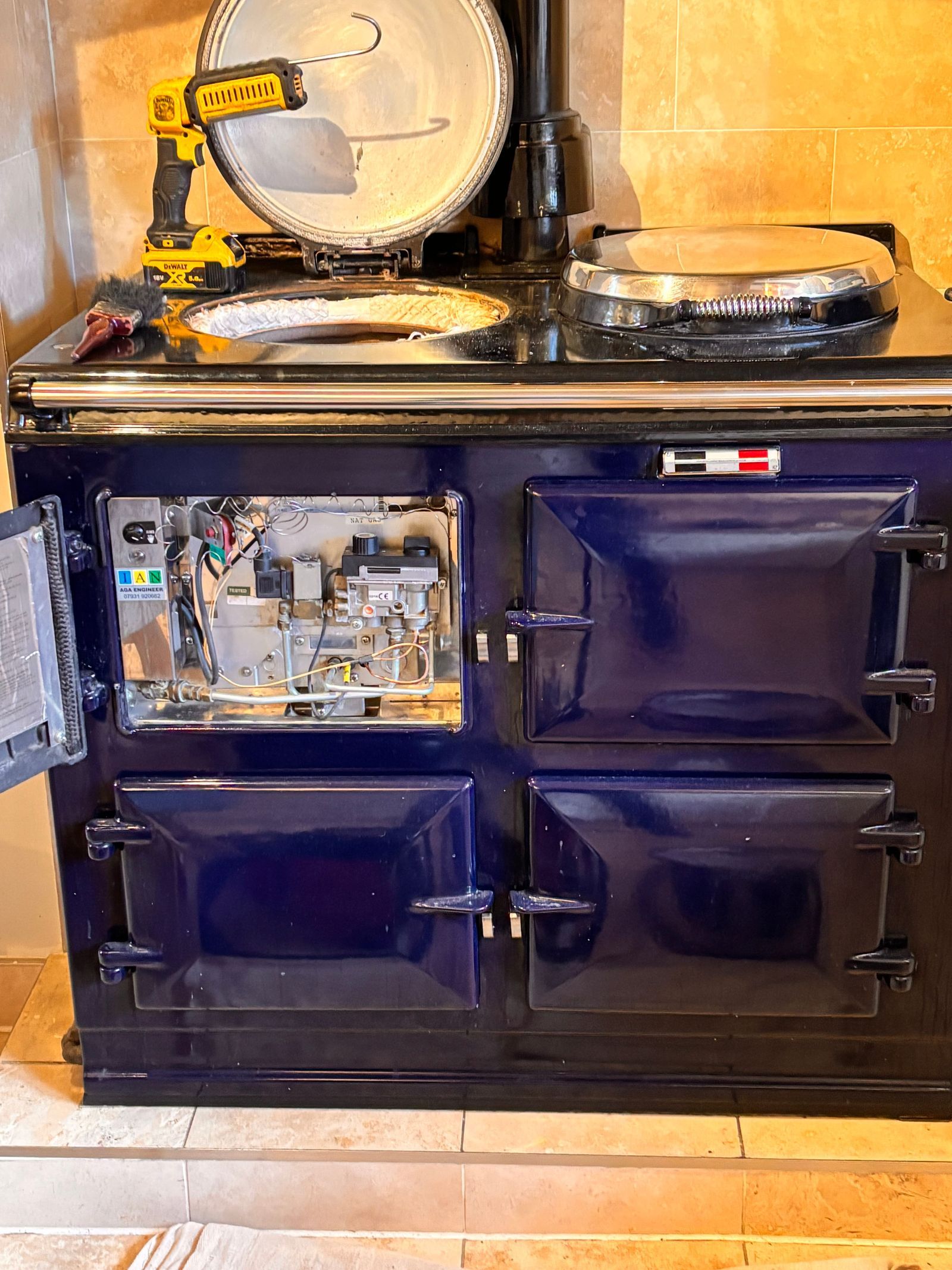 A dark blue three-oven gas AGA with its burner door open to show the gas valve and wiring, and the left hotplate lifted out so the insulation underneath shows
