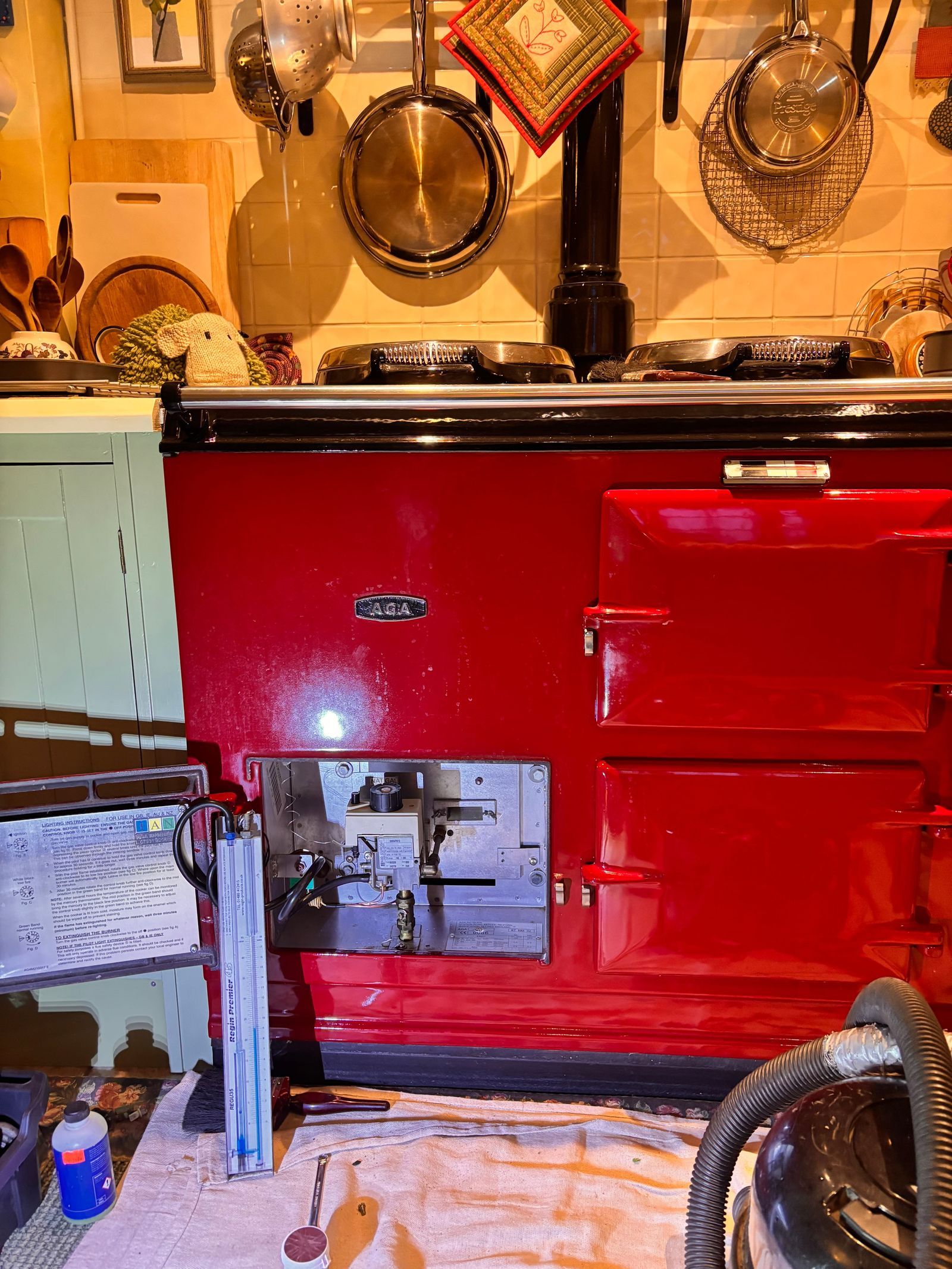 A red two-oven gas AGA with its burner door open to show the gas valve, a pressure gauge standing on the dust sheet in front and pans hanging on the wall above