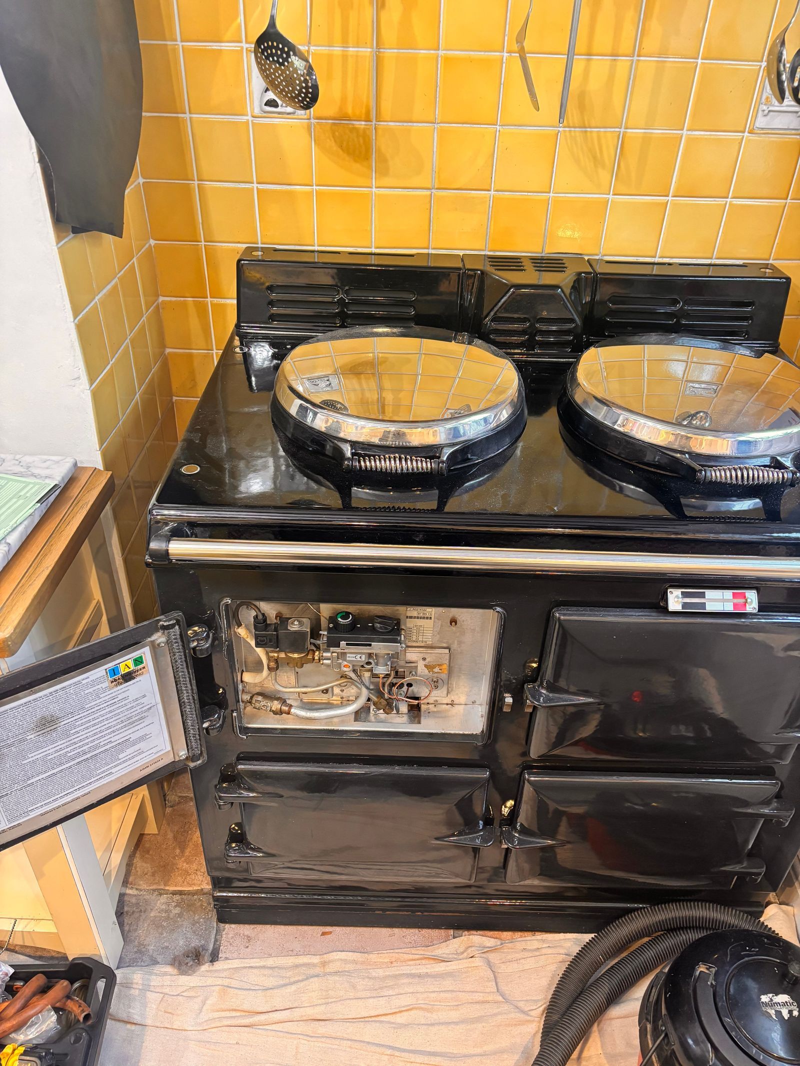 A black gas AGA in a yellow tiled alcove with its burner door open to show the gas valve and controls, a dust sheet on the floor and a vacuum cleaner beside it