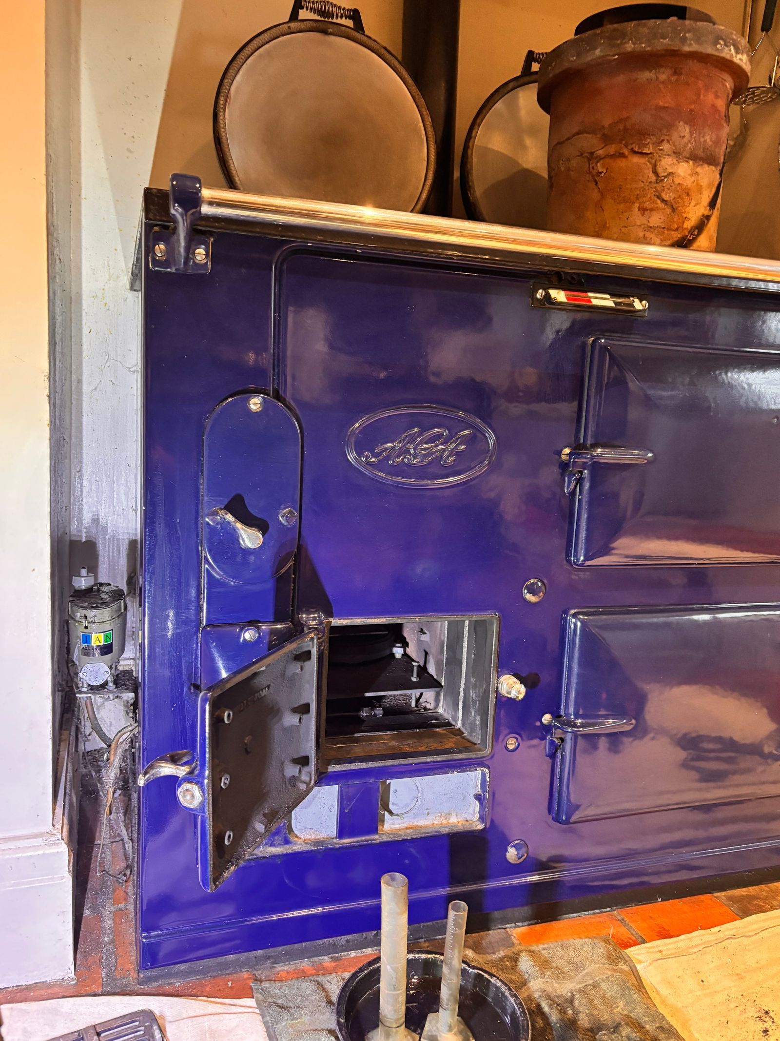 A dark blue oil AGA with its lower front door open to show the burner, the oil control valve on the wall to its left, and burner parts on a dust sheet in front