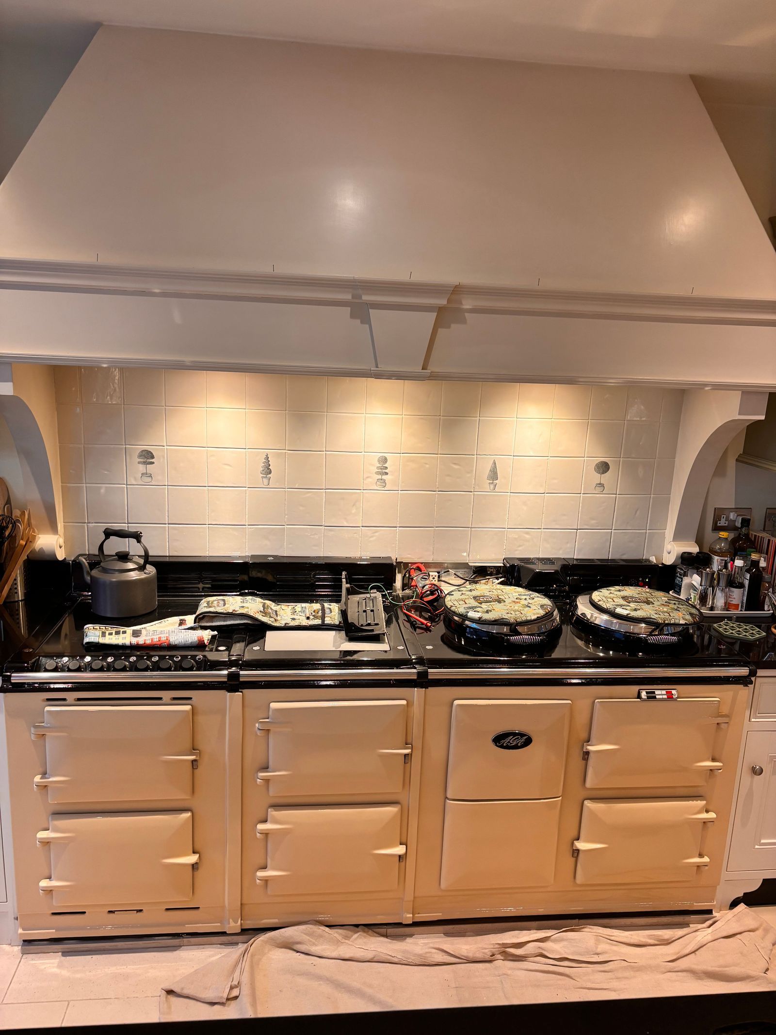 A long cream electric AGA under a cream cooker hood, with a meter and test leads on the top plate and a dust sheet on the floor