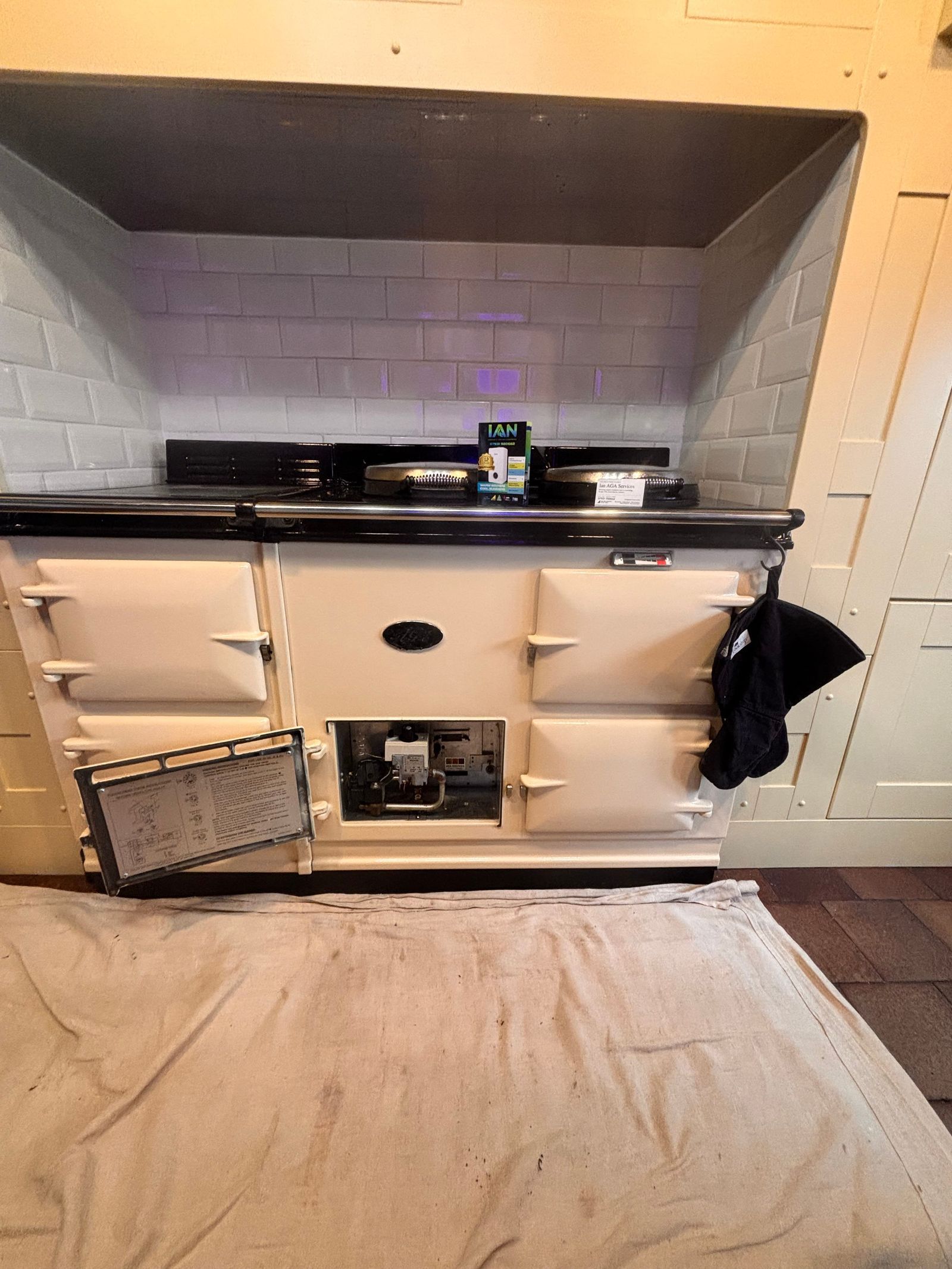 A cream gas AGA in a white tiled alcove with its burner door open to show the gas valve and controls, and a dust sheet on the floor in front