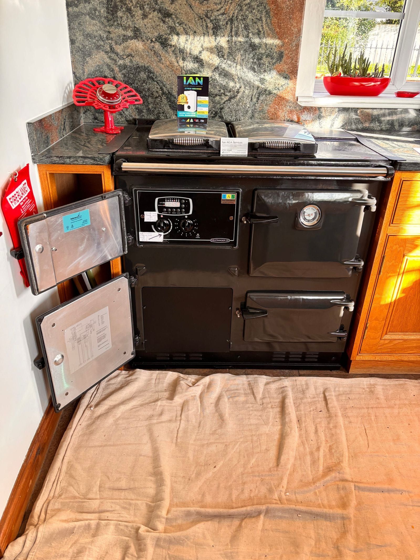 A black gas Rayburn Heatranger CD with its two left-hand doors open, set between granite worktops and oak units, with a dust sheet on the floor in front